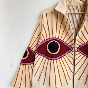 Puede incluir: Chaqueta beige con dise&ntilde;os de ojos bordados. La chaqueta tiene cuello, cierre de cremallera y mangas largas. Los dise&ntilde;os de ojos son en burdeos, rosa y negro, con l&iacute;neas radiantes marrones. El forro interior tiene un estampado de rayas.