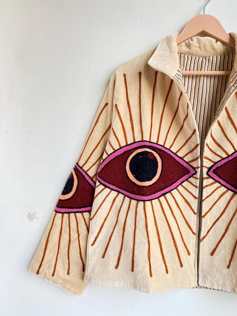 Puede incluir: Chaqueta beige con dise&ntilde;os de ojos bordados. La chaqueta tiene cuello, cierre de cremallera y mangas largas. Los dise&ntilde;os de ojos son en burdeos, rosa y negro, con l&iacute;neas radiantes marrones. El forro interior tiene un estampado de rayas.