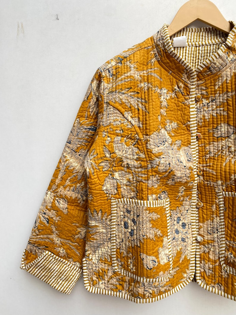 May include: A mustard yellow quilted jacket with a floral print and white piping. The jacket has a mandarin collar, a button closure, and two patch pockets.