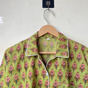 May include: Green pajama top with a floral pattern in pink and orange. The top has white trim, a chest pocket, and is button-down. The garment is hanging on a wooden hanger.