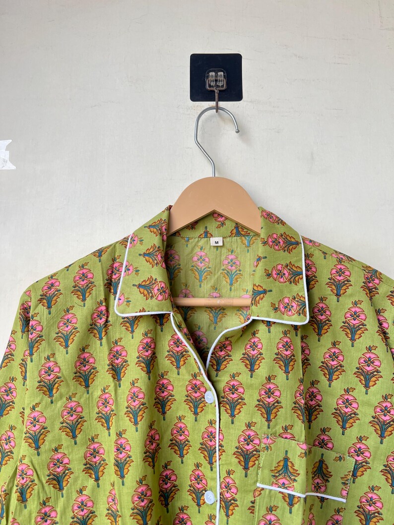 May include: Green pajama top with a floral pattern in pink and orange. The top has white trim, a chest pocket, and is button-down. The garment is hanging on a wooden hanger.