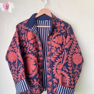 May include: A navy blue jacket with intricate coral-colored floral embroidery. The jacket features a striped lining and cuffs, and is displayed on a wooden hanger. The design includes a collar and long sleeves.