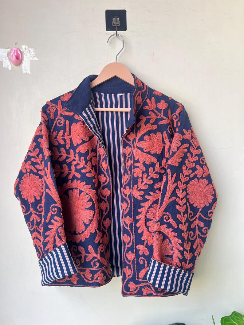 May include: A navy blue jacket with intricate coral-colored floral embroidery. The jacket features a striped lining and cuffs, and is displayed on a wooden hanger. The design includes a collar and long sleeves.