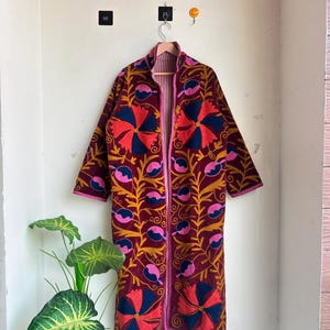 May include: A long-sleeved, open-front coat in a deep burgundy fabric featuring vibrant, large-scale floral embroidery in shades of bright orange, dark navy blue, and pink. The garment has a high collar and is finished with pink piping along the edges.