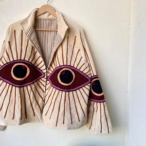 Puede incluir: Chaqueta beige con dise&ntilde;os de ojos bordados. La chaqueta presenta grandes ojos estilizados en tonos burdeos, rosa y negro, con l&iacute;neas radiales. La chaqueta est&aacute; colgada en una percha de madera contra una pared blanca.