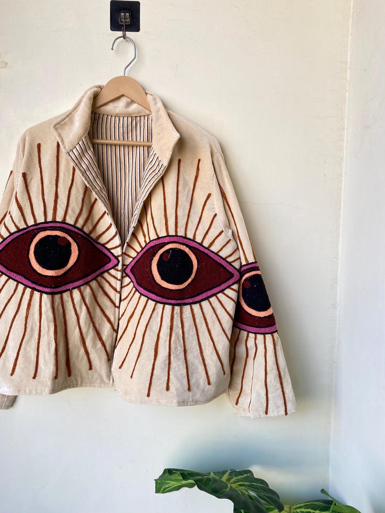Puede incluir: Chaqueta beige con dise&ntilde;os de ojos bordados. La chaqueta presenta grandes ojos estilizados en tonos burdeos, rosa y negro, con l&iacute;neas radiales. La chaqueta est&aacute; colgada en una percha de madera contra una pared blanca.