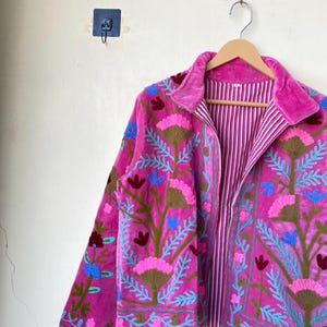 以下が含まれることがあります： ピンク、青、茶色の花の刺繍が施されたマゼンタ色のジャケット。 ジャケットはベルベットの襟とストライプの裏地が付いています。 白い壁を背景に、木製のハンガーに吊るされています。