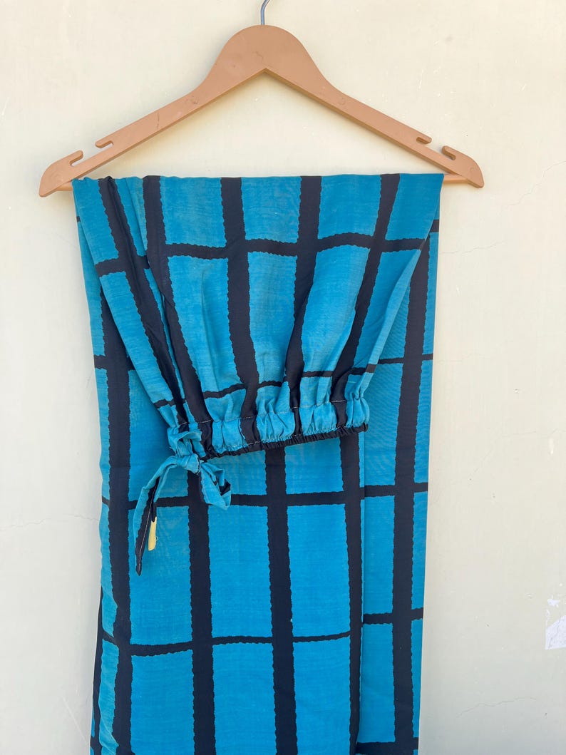 May include: A pair of teal and black patterned pants hanging on a wooden hanger. The pants feature a grid pattern in black over a teal background. The pants have an elastic cuff at the ankle and a drawstring.