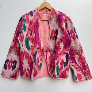 May include: A quilted jacket with a vibrant, abstract pattern in shades of pink, purple, and green. The jacket has a mandarin collar and a button closure. The lining is a solid pink, and the jacket is hanging on a wooden hanger.