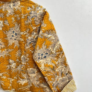 May include: A mustard yellow quilted jacket with a floral print. The jacket has a mandarin collar and long sleeves with striped cuffs.