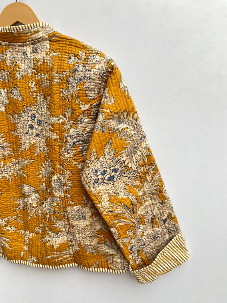 May include: A mustard yellow quilted jacket with a floral print. The jacket has a mandarin collar and long sleeves with striped cuffs.
