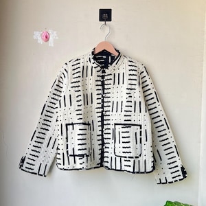 May include: A cream-colored jacket with a black geometric pattern. The jacket has a stand-up collar, two front pockets, and black trim. It is hanging on a wooden hanger against a neutral wall.