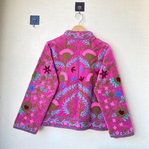 以下が含まれることがあります： 青、緑、茶色の刺繍が施された鮮やかなピンク色のジャケット。 ジャケットはハイカラーと長袖で、詳細なパターンを示しています。 無地の壁に木製のハンガーに吊るされた衣服。