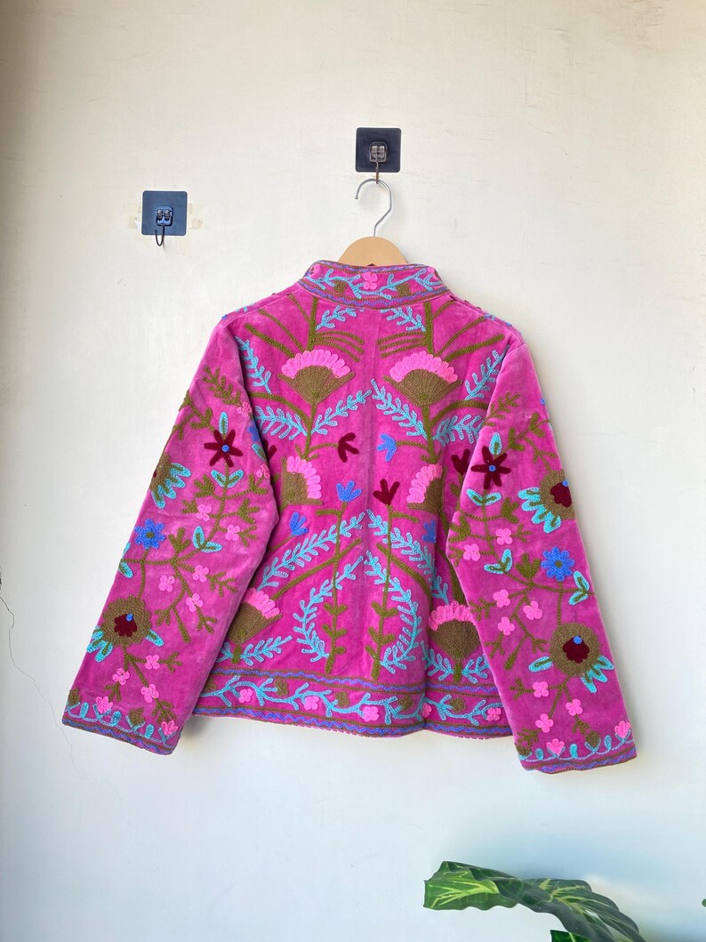 以下が含まれることがあります： 青、緑、茶色の刺繍が施された鮮やかなピンク色のジャケット。 ジャケットはハイカラーと長袖で、詳細なパターンを示しています。 無地の壁に木製のハンガーに吊るされた衣服。