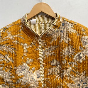 May include: A mustard yellow quilted jacket with a floral print and white and brown striped trim. The jacket has a mandarin collar and a button closure.