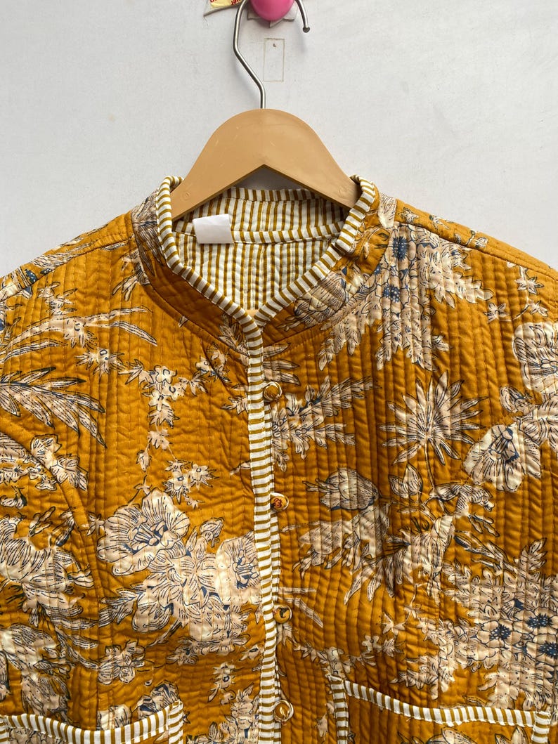May include: A mustard yellow quilted jacket with a floral print and white and brown striped trim. The jacket has a mandarin collar and a button closure.