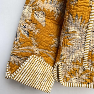 May include: A mustard yellow quilted jacket with a white and blue floral print. The jacket has a striped cuff detail.