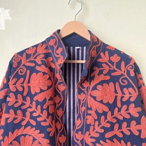 May include: A navy blue jacket with intricate coral-colored floral embroidery. The jacket features a mandarin collar and a striped lining. The embroidery creates a striking contrast against the dark blue fabric, showcasing a detailed floral pattern.