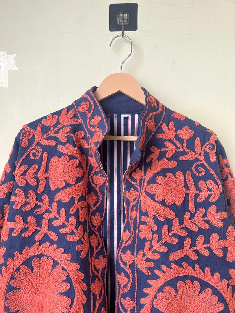 May include: A navy blue jacket with intricate coral-colored floral embroidery. The jacket features a mandarin collar and a striped lining. The embroidery creates a striking contrast against the dark blue fabric, showcasing a detailed floral pattern.