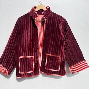 Puede incluir: Una chaqueta acolchada de color burdeos con ribetes de rayas rojas y blancas. La chaqueta tiene dos bolsillos y una cremallera.