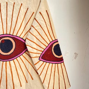 Puede incluir: Chaqueta color crema con dise&ntilde;o de ojo bordado. La chaqueta presenta un gran dise&ntilde;o de ojo bordado en la espalda y la manga, con una pupila azul oscuro, un iris granate y un contorno rosa. El ojo est&aacute; rodeado de rayos bordados en naranja.
