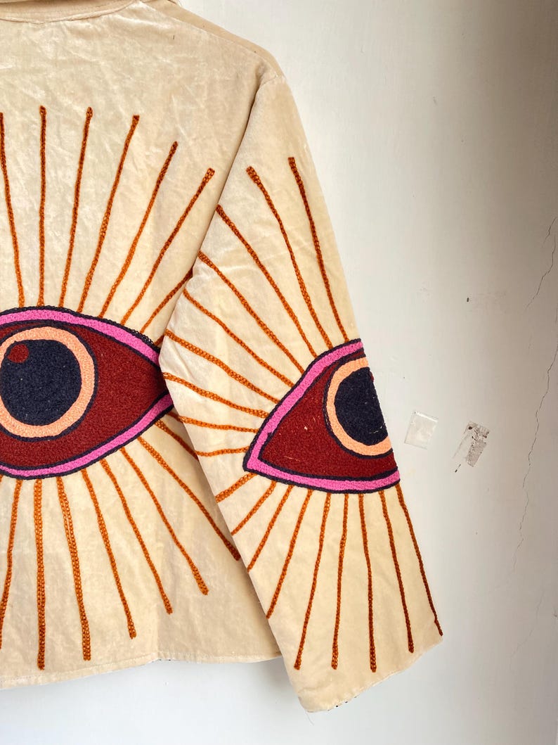 Puede incluir: Chaqueta color crema con dise&ntilde;o de ojo bordado. La chaqueta presenta un gran dise&ntilde;o de ojo bordado en la espalda y la manga, con una pupila azul oscuro, un iris granate y un contorno rosa. El ojo est&aacute; rodeado de rayos bordados en naranja.