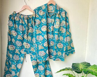 May include: A teal pajama set with a floral pattern, including a button-down shirt and drawstring pants. The set features white trim and is hanging on a wooden hanger. The floral design includes white and light green flowers.
