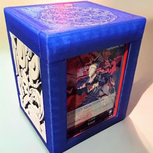 May include: A blue, cube-shaped card holder with a decorative white design on one side. The holder features a clear window displaying a card with a character illustration. The top has a circular pattern. The text "RetroJoe3D" is at the bottom.