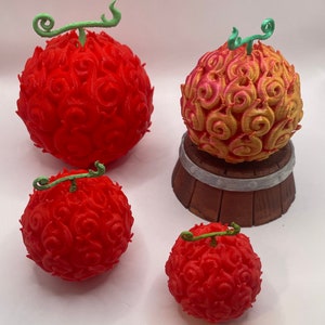 Op de afbeelding: Vier 3D-geprinte fruitmodellen, die lijken op de "Vlam-Vlam-Vrucht" uit de anime "One Piece". Drie zijn rood, één is rood en geel, en ze hebben allemaal groene stelen.