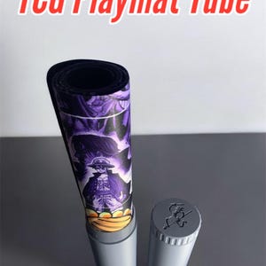 Może przedstawiać: Szary tubus z zwiniętą wewnątrz czarną i fioletową matą do gry. Tubus ma szary korek z logo "RetroJoe3D". Mata do gry ma fioletowo-czarny wzór z postacią w środku.