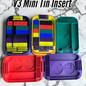 May include: A collection of colorful V3 Mini Tin Inserts. The inserts are in various colors, including blue, yellow, green, red, and purple. Each insert has a unique design with slots and compartments. The text "V3 Mini Tin Insert" is at the top.