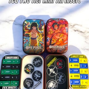 Könnte beinhalten: Zwei TCG Two Tier Mini Tin Inserts mit One Piece-Kartenspiel-Artwork. Enthält schwarz-weiße Token mit Totenkopf-Designs und Fähigkeitszähler in Grün und Gelb.