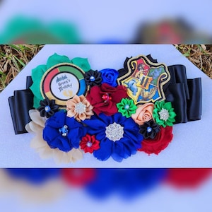 May include: A black satin sash with a Harry Potter themed baby shower decoration. The decoration features a Hogwarts crest, a green and gold circle with the text "Haven's Mommy" and a pair of baby footprints, and a variety of colorful fabric flowers.