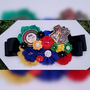 May include: A black sash with a large, gold Hogwarts crest and a smaller circle with the text "It's a Girl" in white. The sash is decorated with colorful fabric flowers in red, yellow, blue, and green.