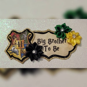 May include: A gold and black Hogwarts house crest with a black, green, and yellow ribbon flower embellishment. The text "Big Brother To Be" is on a parchment-like background.