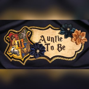 May include: A gold and black Hogwarts house crest with a decorative sign that says "Auntie To Be". The sign is decorated with blue, teal, and beige fabric flowers.