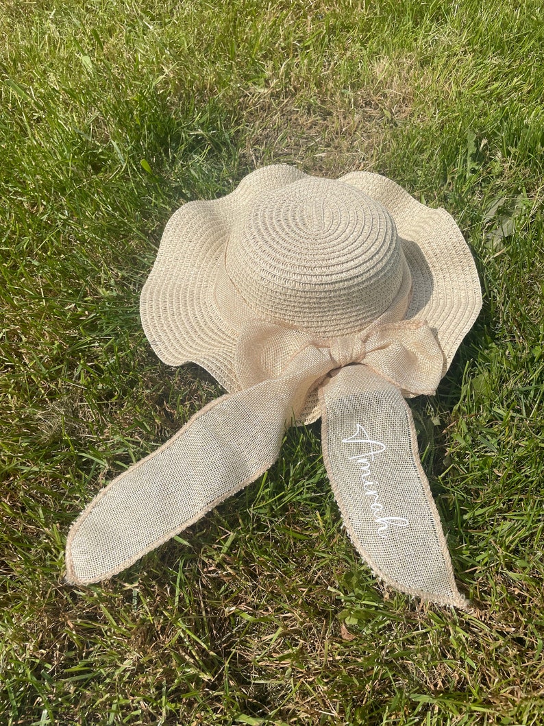 May include: A beige sun hat with a wide, wavy brim and a large bow. The bow is made of a textured, light-colored fabric. The word "Amara" is written in cursive on one of the bow's streamers. The hat is lying on a bed of green grass.