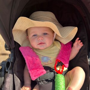 May include: A baby in a stroller wearing a beige sun hat and a yellow onesie with an elephant graphic. The stroller has pink strap covers and a green and white toy. The baby is looking at the camera.