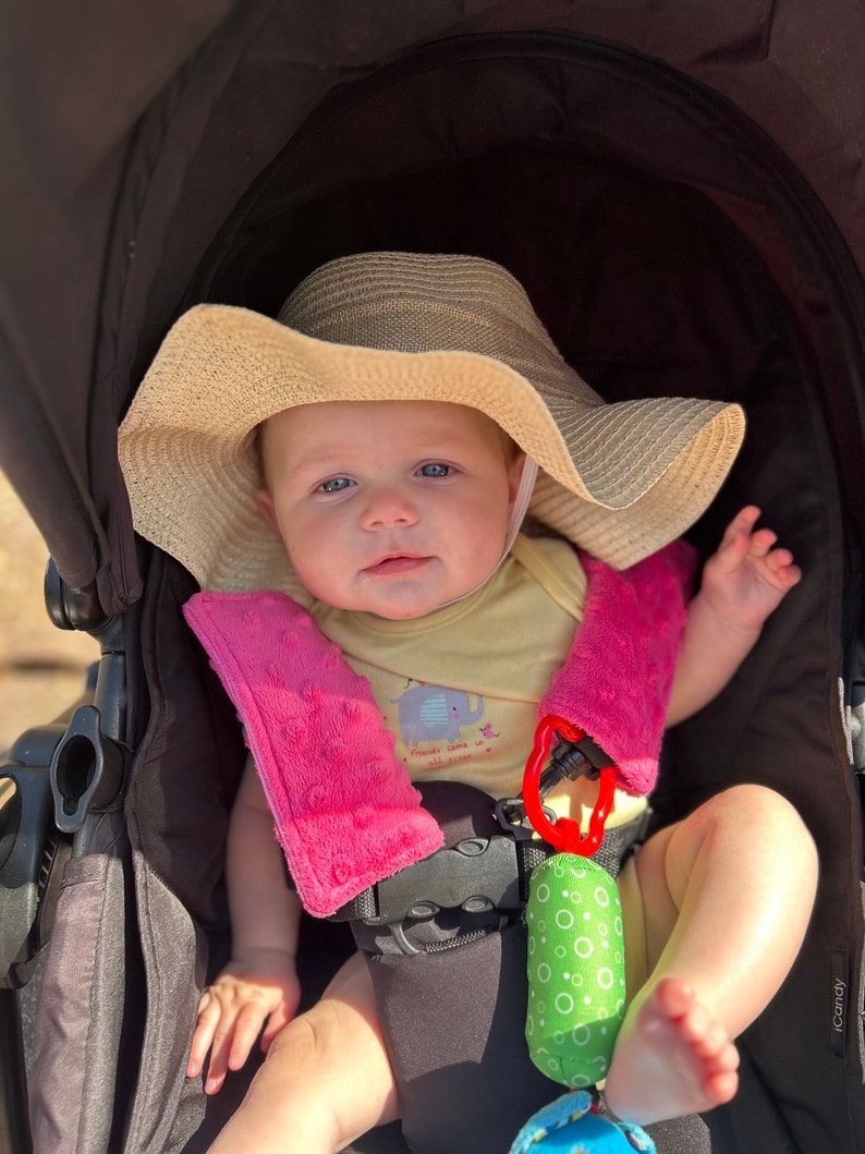 May include: A baby in a stroller wearing a beige sun hat and a yellow onesie with an elephant graphic. The stroller has pink strap covers and a green and white toy. The baby is looking at the camera.