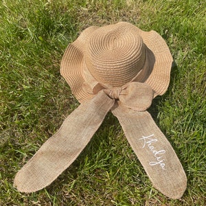 May include: A beige sun hat with a wide brim and a large bow. The hat has long, flowing ribbons with the name "Khadija" in white script. The hat is made of woven straw and is lying on green grass.