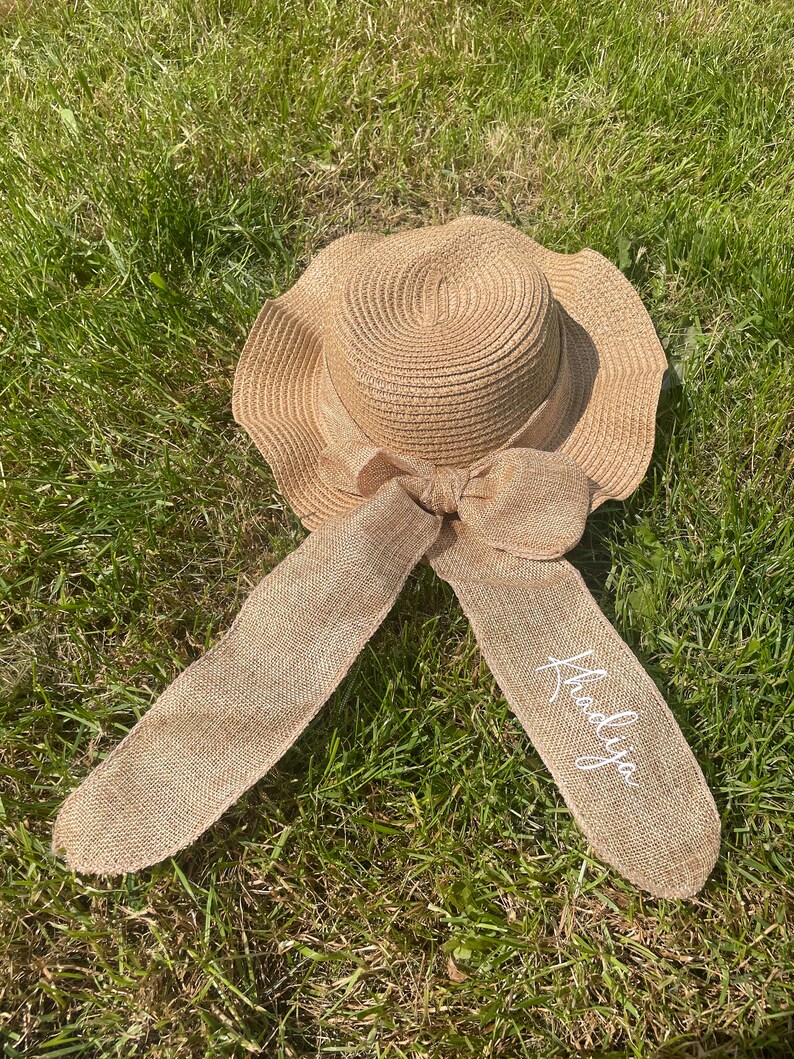 May include: A beige sun hat with a wide brim and a large bow. The hat has long, flowing ribbons with the name "Khadija" in white script. The hat is made of woven straw and is lying on green grass.