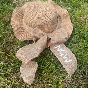 May include: A woven straw sun hat with a wide, wavy brim and a burlap ribbon tied in a bow. The ribbon has the letters "NGW" printed on it in white. The hat is lying on a bed of green grass.