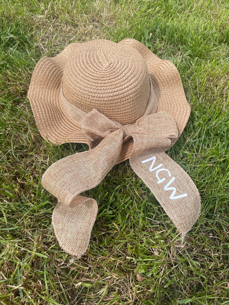 May include: A woven straw sun hat with a wide, wavy brim and a burlap ribbon tied in a bow. The ribbon has the letters "NGW" printed on it in white. The hat is lying on a bed of green grass.