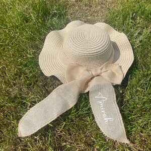 May include: A beige sun hat with a wide, wavy brim and a large bow made of burlap-like material. The bow has long streamers, one of which is embroidered with the name "Amirah" in white script. The hat is lying on green grass.