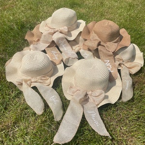 May include: A collection of straw sun hats with wide brims and decorative bows. Each hat has a ribbon with a name embroidered on it. The hats are in shades of beige and brown, resting on green grass.