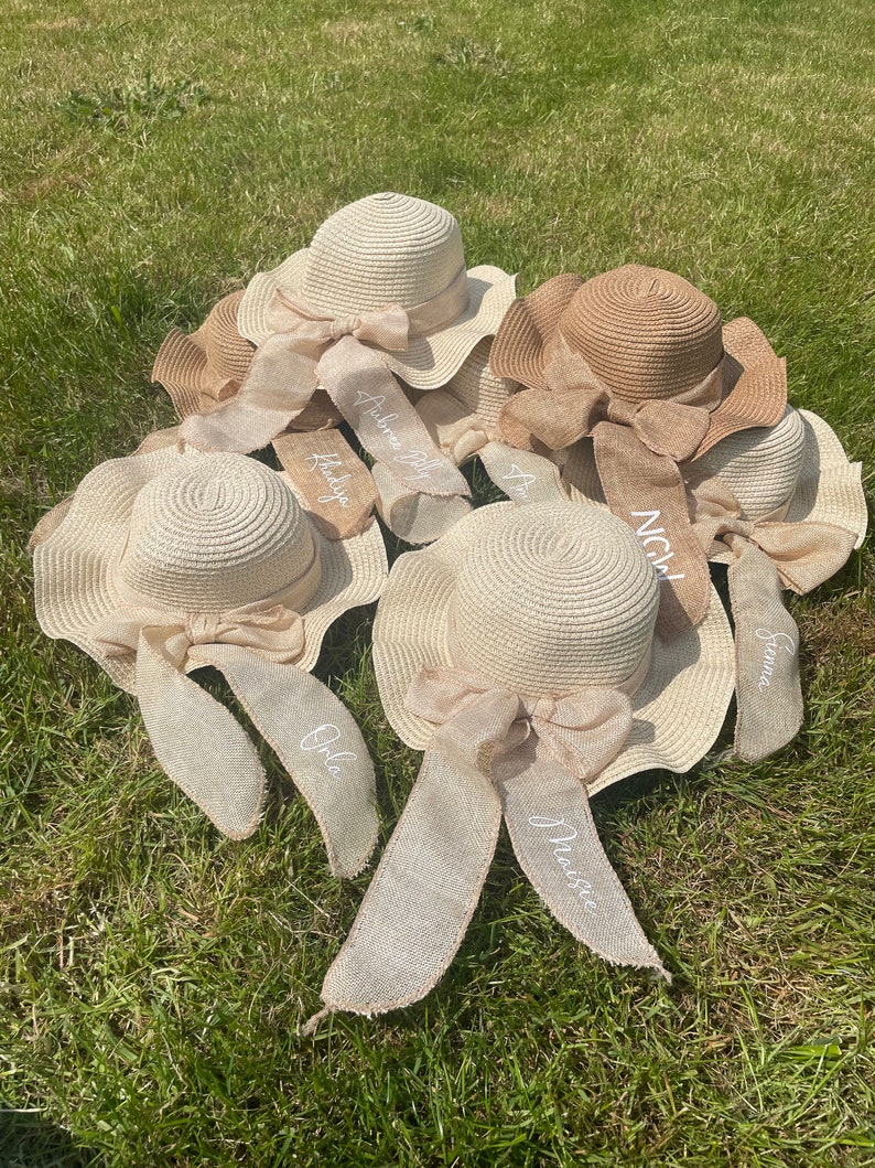 May include: A collection of straw sun hats with wide brims and decorative bows. Each hat has a ribbon with a name embroidered on it. The hats are in shades of beige and brown, resting on green grass.