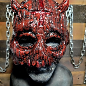 May include: A red devil mask with horns and a cross painted on the forehead. The mask is made of a textured material and has a realistic look.