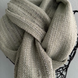 May include: A woven scarf in a light sage green and off-white color palette. The fabric features a textured, cross-hatched pattern with visible interlaced threads. The scarf is draped in a loose knot, displaying the consistent weave throughout.