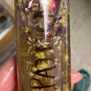 May include: A clear glass bottle filled with a golden liquid and various dried flowers and herbs. The bottle is labelled "PALA".