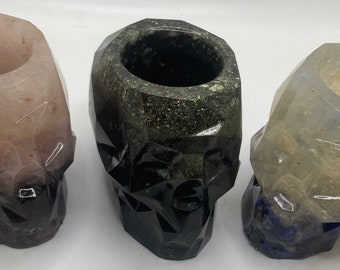 Puede incluir: Tres portavelas de calavera de cristal, uno es rosa, otro es negro y otro es transparente con detalles azules.